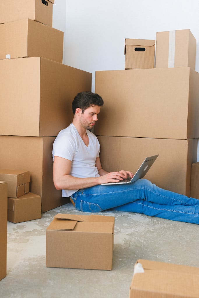 Young man using a laptop surrounded by stacked moving boxes indoors, depicting multitasking during relocation.